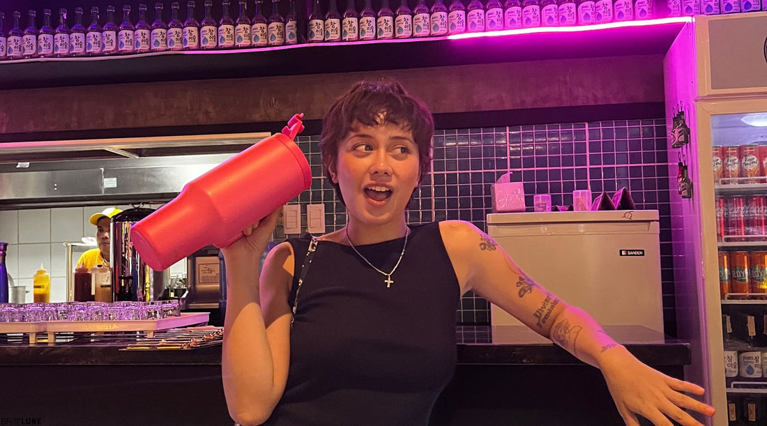 A person in a dark outfit holds a vibrant pink drink container, set against a backdrop of colorful bottles in a lively bar atmosphere.